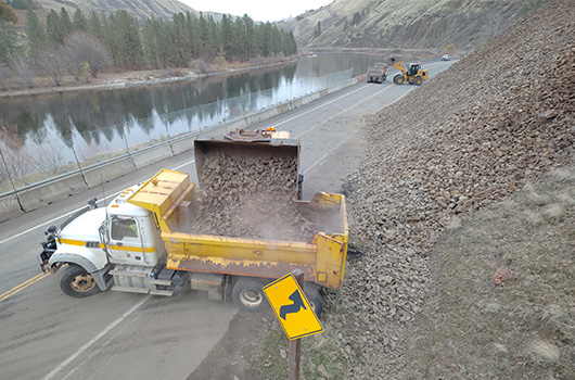 On the US-12 emergency slope project near Arrow, a loader empties a bucket of rocky debris over a truck bed.