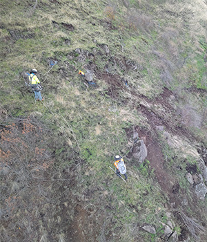 Workers attached to ropes carefully transverse a rocky slope high above US-12 by Arrow bridge.