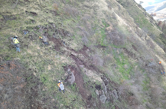 Workers attached to ropes carefully transverse a rocky slope high above US-12 by Arrow bridge.