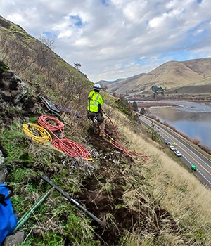 Picture of a worker on the slope above US-12 near Arrow Bridge. He is pulling on a line and the river can be seen in the background.