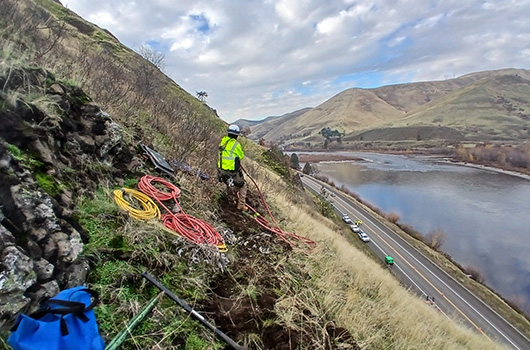 High up on the slope above US-12 near Arrow Bridge, a rock scaling worker pulls on a line.