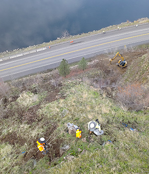 Workers can be seen on the slope above US-12 and equipment excavates in the distance.