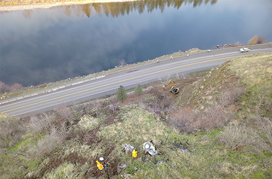 Workers can be seen on the slope above US-12 and equipment excavates in the distance.
