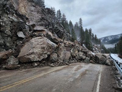 Rockslide debris covering part of a two-lane mountain highway, with large boulders at the base of a rocky hillside.