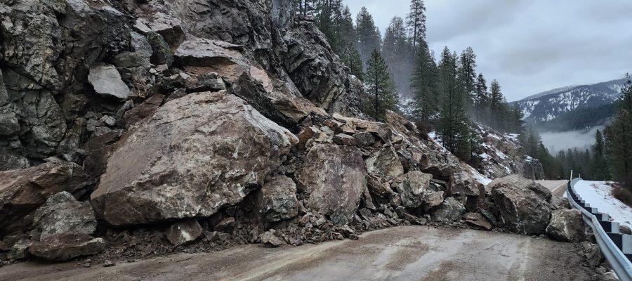 A landslide has sent multiple large boulders and smaller rock fragments from a steep, exposed rock slope onto a two-lane mountain highway. The debris fully blocks the roadway near a guardrail. Pine trees line the slope and surrounding hillsides, with patches of snow visible under an overcast sky.