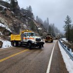 Maintenance crews using heavy equipment to clear rockslide debris from a mountain highway with snow along the roadside.