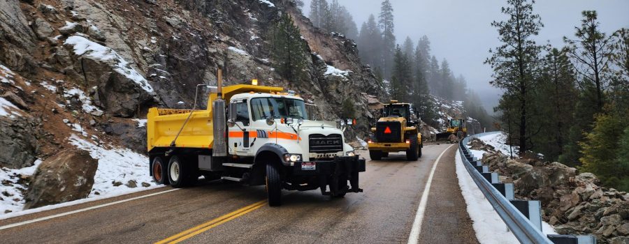 Maintenance crews using heavy equipment to clear rockslide debris from a mountain highway with snow along the roadside.