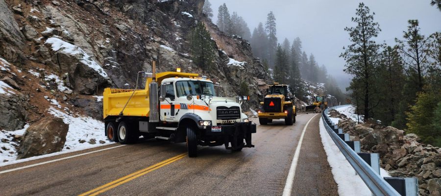 Maintenance crews using heavy equipment to clear rockslide debris from a mountain highway with snow along the roadside.