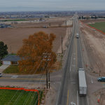 Aerial view of State Highway 55 at Montana Avenue, showing surrounding farmland, nearby homes, utility lines, and traffic at the intersection. One side of the roadway shows cleared ground for upcoming construction.