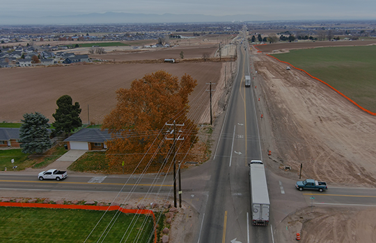 Aerial view of State Highway 55 at Montana Avenue, showing surrounding farmland, nearby homes, utility lines, and traffic at the intersection. One side of the roadway shows cleared ground for upcoming construction.