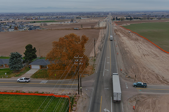 Aerial view of State Highway 55 at Montana Avenue, showing surrounding farmland, nearby homes, utility lines, and traffic at the intersection. One side of the roadway shows cleared ground for upcoming construction.