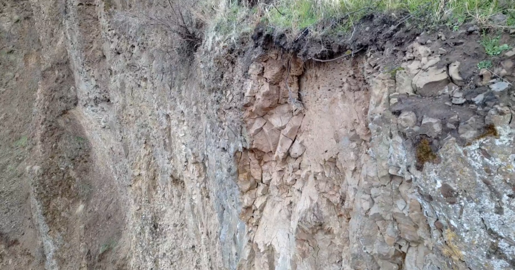 Figure 3. Loose rock identified as a hazard by geologist awaiting removal by safety scaling crew. Green vegetation can be seen near the top of the picture and the remainder is a large vertical slope composed of dark colored rocks.
