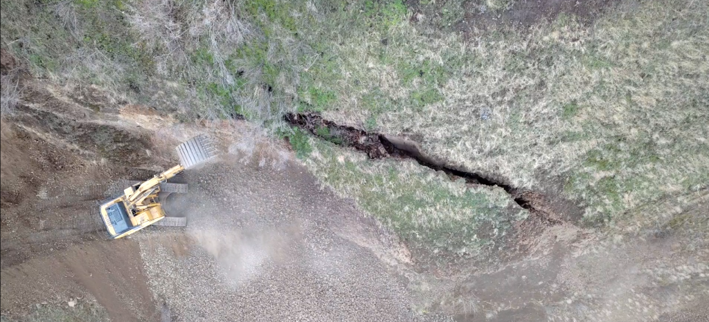 Excavator removing the area that began to destabilize above the pad during scaling as directed by geologist piloting the drone. The fissure can be seen in the vegetation above and the second half of the photo below shows the equipment on the rocky slope.