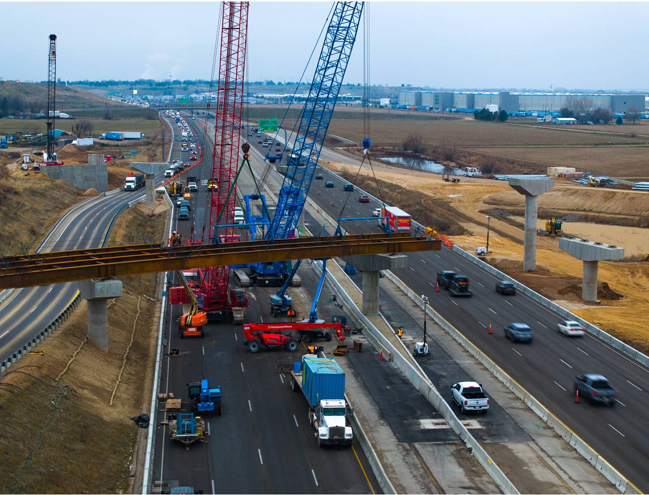 Horizontal girders arch across a busy six-lane highway as cranes overlook the area.