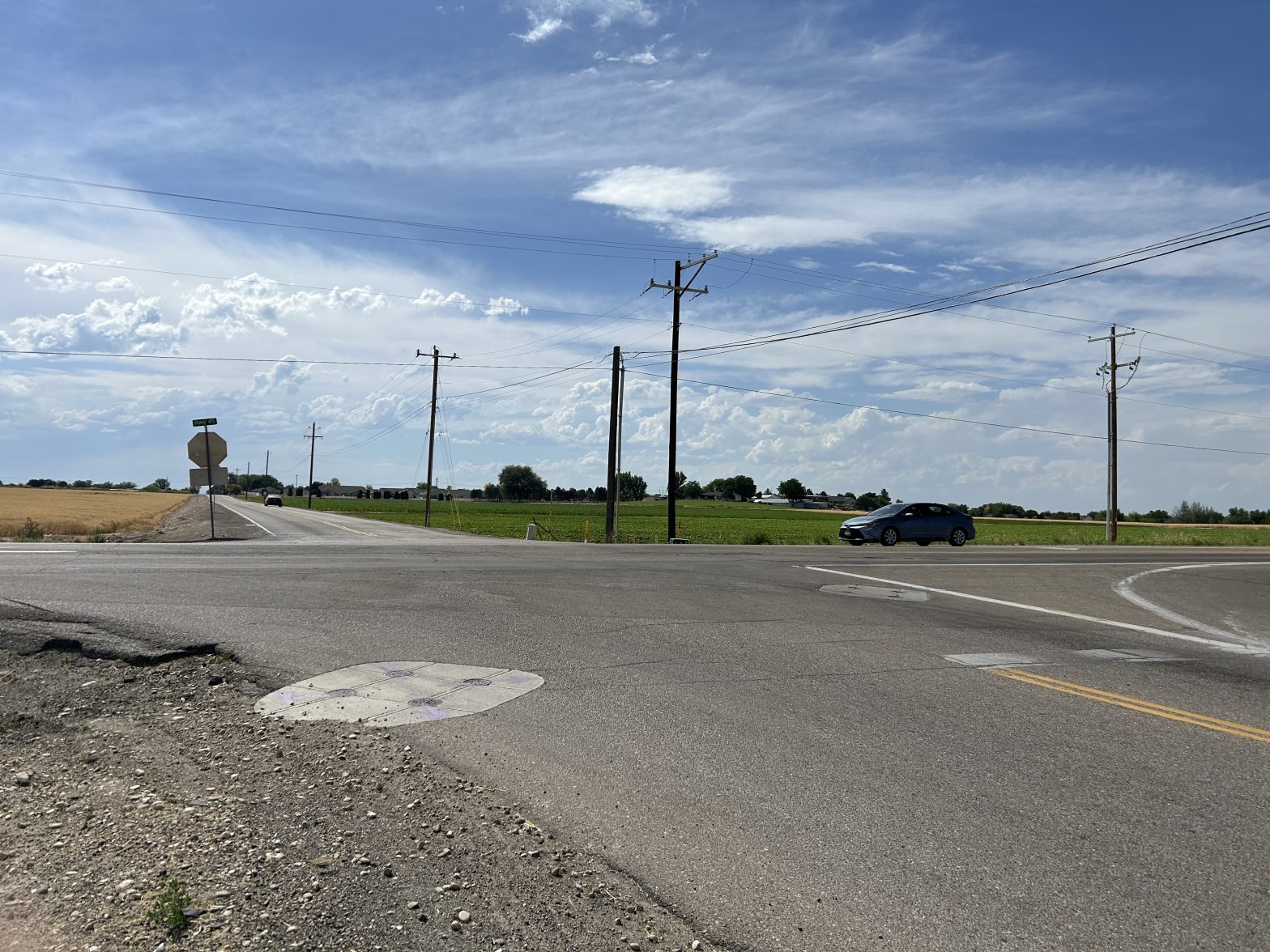 Since car pulls up to intersection with stop sign visible. Fields, blue sky with white clouds in background.