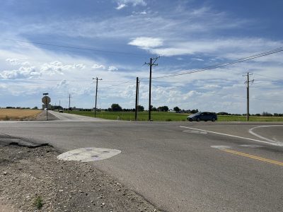 Single car pulls up to intersection with stop sign visible. Fields, blue sky with white clouds in background.
