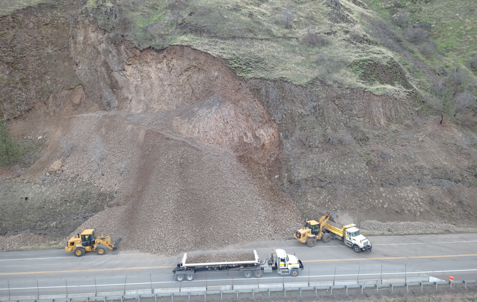 Photograph taken during slope assessment following scaling and prior to reopening the road. Loaders in the foreground are filling up dump trucks with large buckets full of rocky debris.