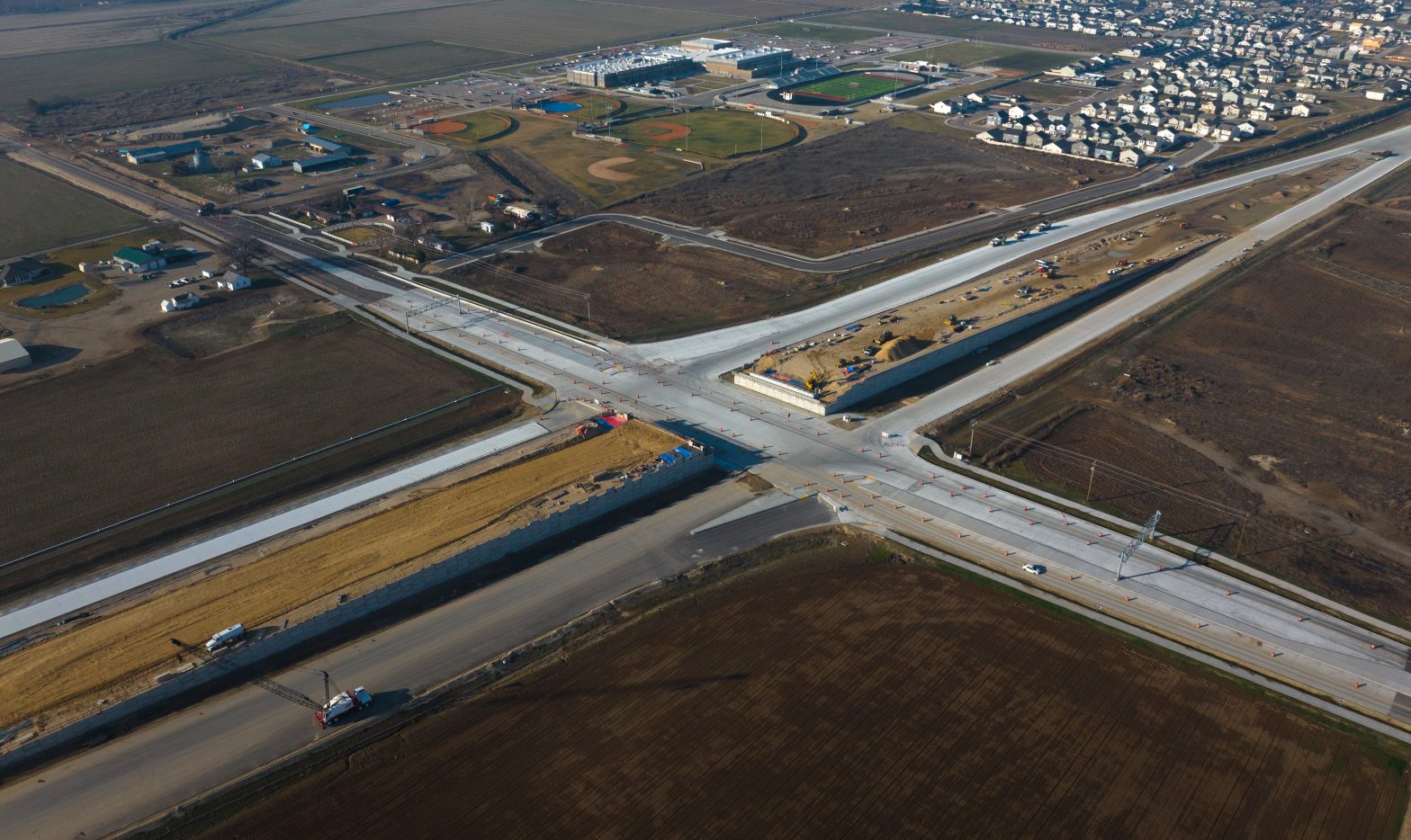 Aerial view of a highway construction site showing a newly built overpass and widened roadway at a four-way intersection, with concrete lanes, traffic cones, construction equipment, and surrounding farmland and residential neighborhoods.
