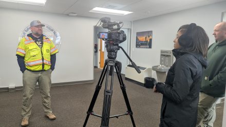 Man in high visibility jacket stands in front of a media camera while a reporter stands behind camera performing interview.