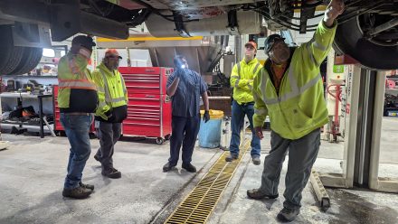 Five transportation workers in high-visibility jackets stand beneath a raised vehicle in a maintenance shop, observing and discussing components during a hands-on training session.