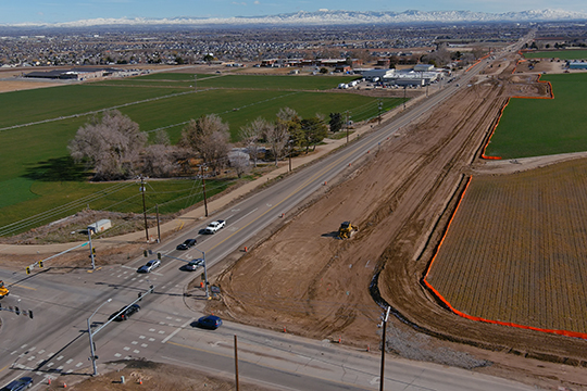 Aerial view of SH-55 construction showing a widened roadway corridor, active earthwork, and an intersection with traffic traveling through the work zone, with surrounding farmland and distant foothills.