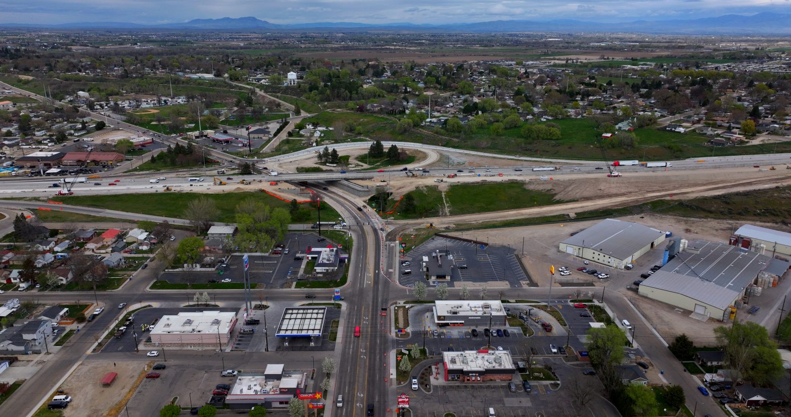 Aerial view of the I-84 interchange area in Nampa, showing ongoing construction with an overpass bridge, earthwork, and traffic moving through the work zone. Surrounding neighborhoods, businesses and open land are visible, with mountains in the distance.