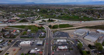 Aerial view of the I-84 interchange area in Nampa, showing ongoing construction with an overpass bridge, earthwork, and traffic moving through the work zone. Surrounding neighborhoods, businesses and open land are visible, with mountains in the distance.