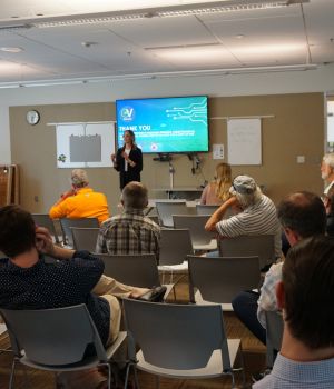 Presenter speaking to a seated audience at the 2022 Idaho NEVI public meeting in Boise, with a presentation slide displayed on a screen.