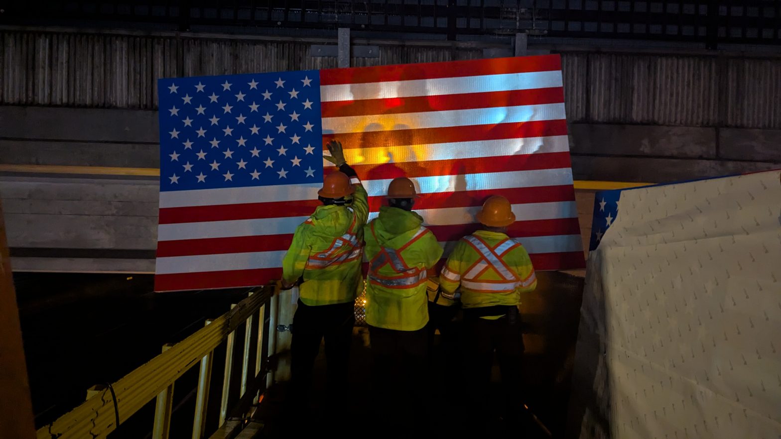ITD workers in high-visibility jackets and hard hats install a large American flag sign on an overpass at night, illuminated by work lights.