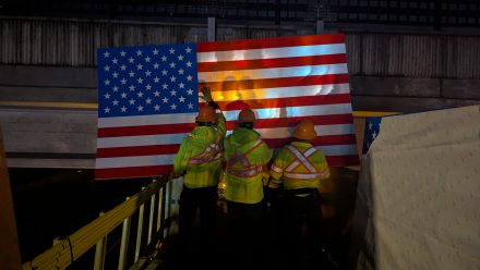 ITD workers in high-visibility jackets and hard hats install a large American flag sign on an overpass at night, illuminated by work lights.