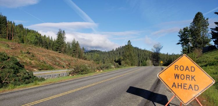 A rural highway curves through a forested area with freshly cut trees along the roadside. An orange “Road Work Ahead” sign stands in the foreground.
