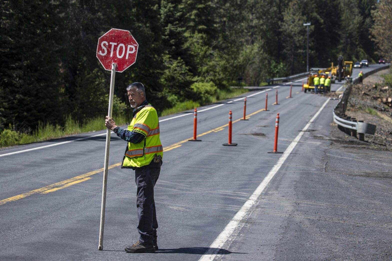 Man in yellow safety vest holds a stop sign while a crew works behind orange candlesticks in a work zone on the right side of the highway.