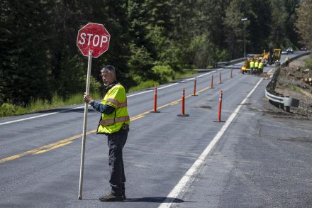 Man in yellow safety vest holds a stop sign while a crew works behind orange candlesticks in a work zone on the right side of the highway.