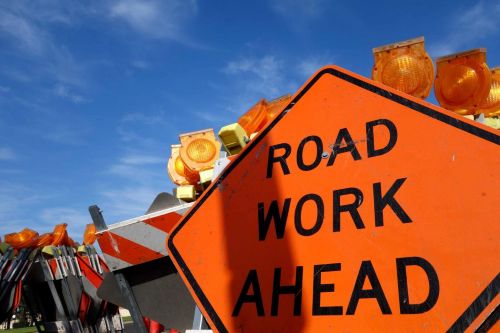 A close-up of an orange “Road Work Ahead” sign with flashing amber lights against a blue sky.