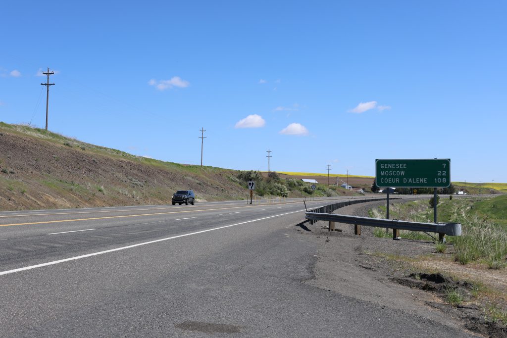 A highway stretches through rolling hillsides with a green road sign showing distances to Genesee, Moscow and Coeur d’Alene.