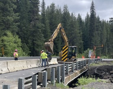 Workers and an excavator perform repairs on a bridge along State Highway 11, with temporary concrete barriers and guardrail visible in a forested setting.