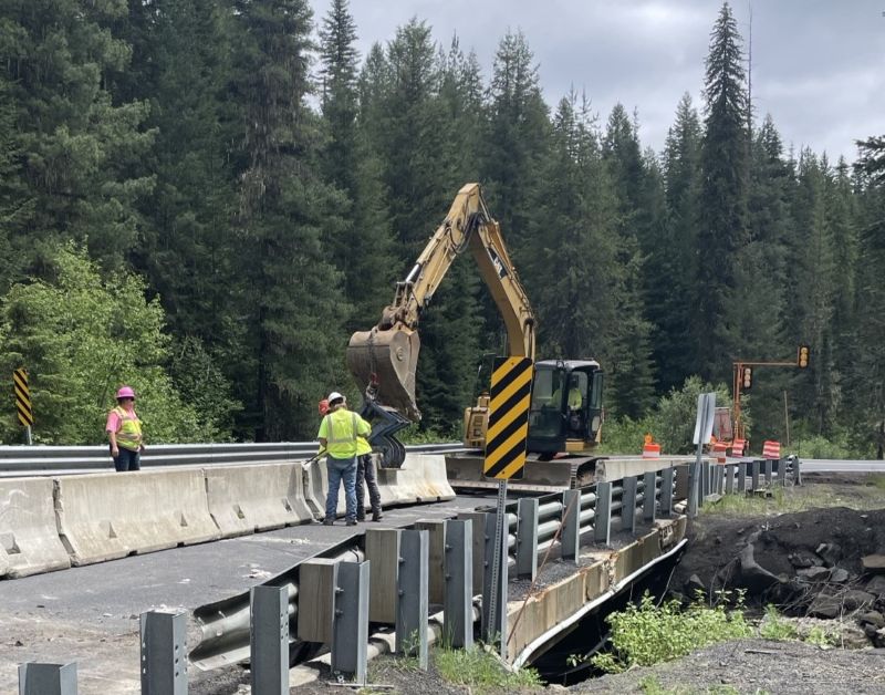 Workers and an excavator perform repairs on a bridge along State Highway 11, with temporary concrete barriers and guardrail visible in a forested setting.