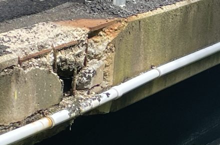 Close-up of bridge damage showing deteriorated concrete, exposed reinforcing steel and corrosion along the edge of the Quartz Creek Bridge above the creek below.