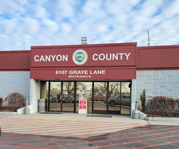 Canyon County building entrance with red-and-white exterior, county seal, and glass double doors under signage reading “Canyon County” and “6107 Graye Lane.”