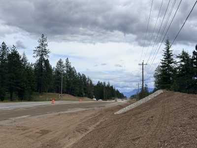 Raodside view of a highway under construction with orange traffic barrels dividing the lanes, gravel shoulders, and dirt piles at the edge of the road.