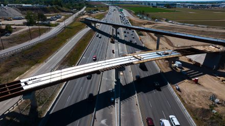 Aerial view of eastbound Interstate 84 showing active traffic traveling beneath two bridge structures under construction. Steel girders span the interstate while crews continue building the new overpasses alongside moving traffic.