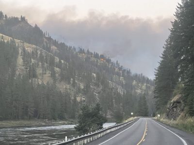 A two-lane highway follows a river through a forested canyon as smoke rises from an active wildfire burning on the hillside above. Flames are visible among the trees while the roadway remains clear in the foreground.