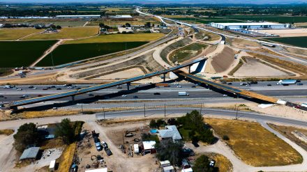 Aerial view of the I-84 and SH-16 interchange under construction, with new elevated ramps crossing the interstate as traffic moves below. Graded roadways, bridge structures, construction equipment, farmland and nearby industrial buildings surround the work zone.