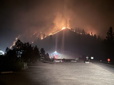 Wildfire burns along a forested ridgeline at night, with flames and heavy smoke visible above a highway where emergency crews and vehicles are staged.