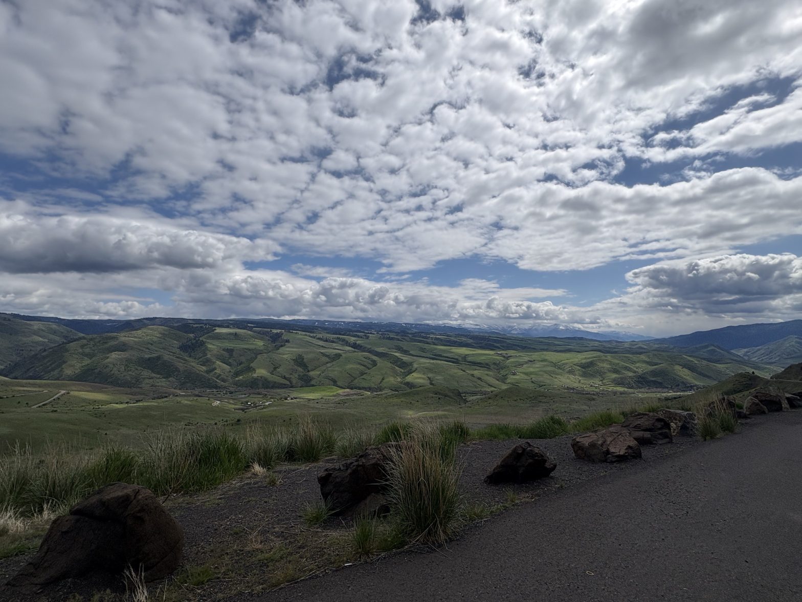 Panoramic view of rolling green hills and distant mountains beneath a dramatic sky filled with layered white and gray clouds. Large rocks and grasses line the edge of the overlook in the foreground.