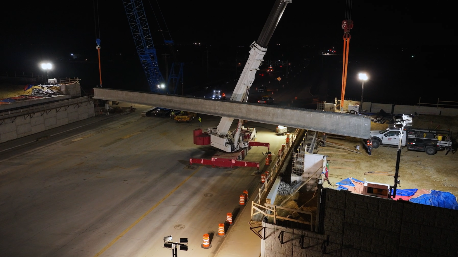 Crews use two cranes to lift a large concrete girder into place over a roadway at night, with the work area illuminated by portable lights.