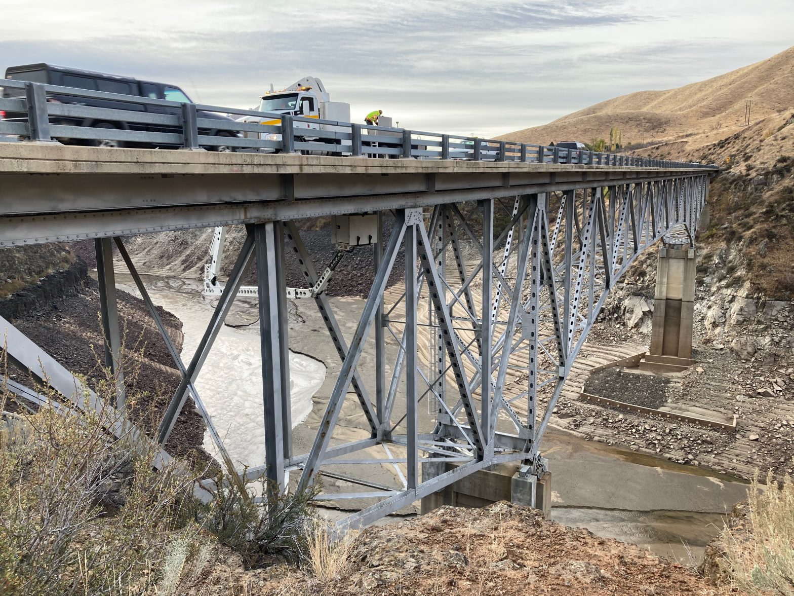 Vehicles travel across the SH-21 Mores Creek Bridge above Mores Creek, with the steel truss structure visible beneath the bridge deck.
