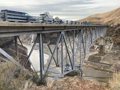 Vehicles travel across the SH-21 Mores Creek Bridge above Mores Creek, with the steel truss structure visible beneath the bridge deck.