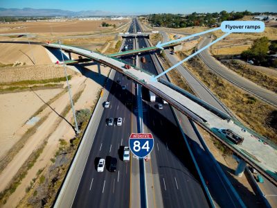 Aerial view of the SH-16 and I-84 interchange showing two flyover ramps under construction above traffic on I-84. Arrows identify the elevated flyover ramps crossing the interstate.