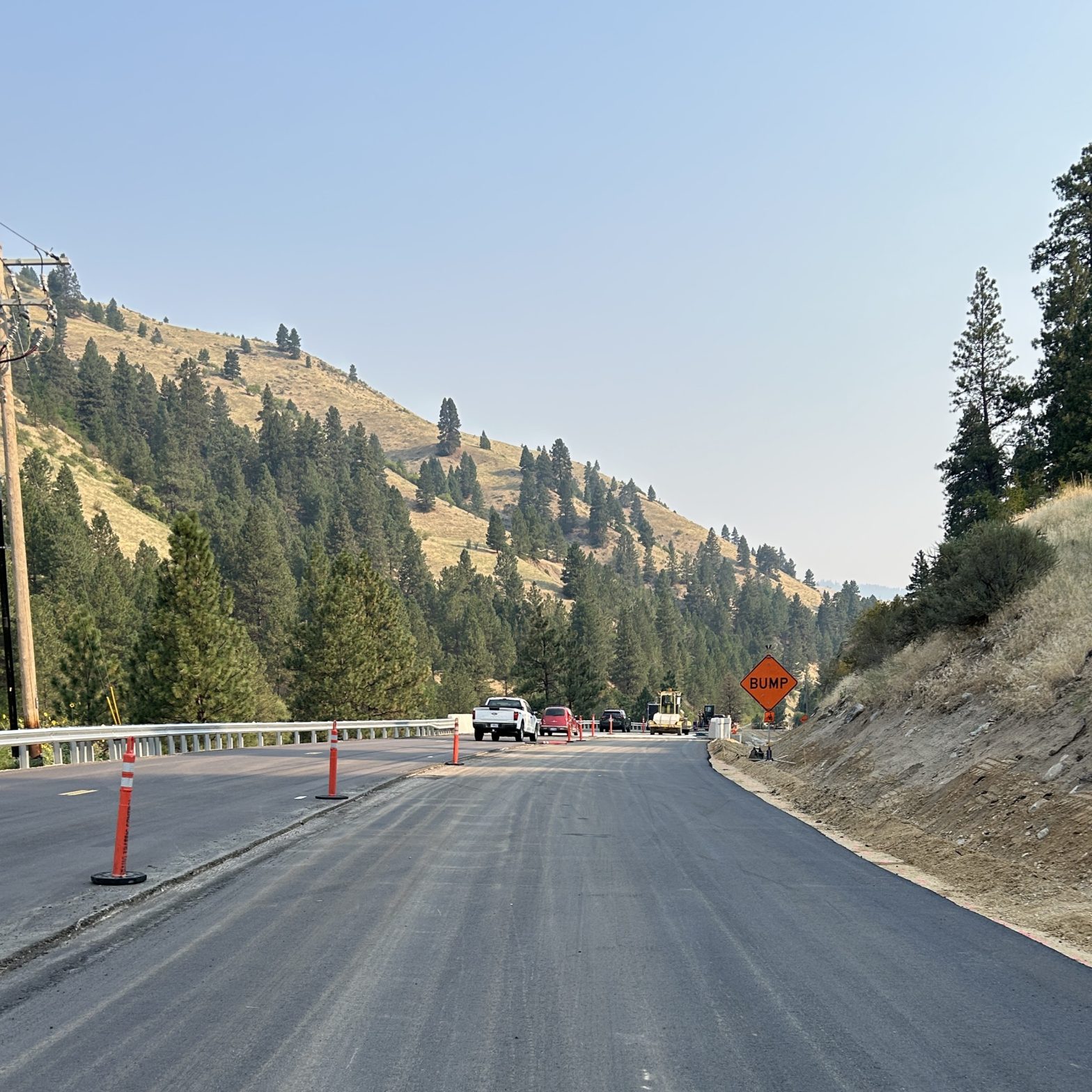 Fresh pavement on SH-55 near the Fleming Creek Bridge, with traffic cones, construction vehicles and a “Bump” warning sign marking the active work zone in a mountainous, forested area.